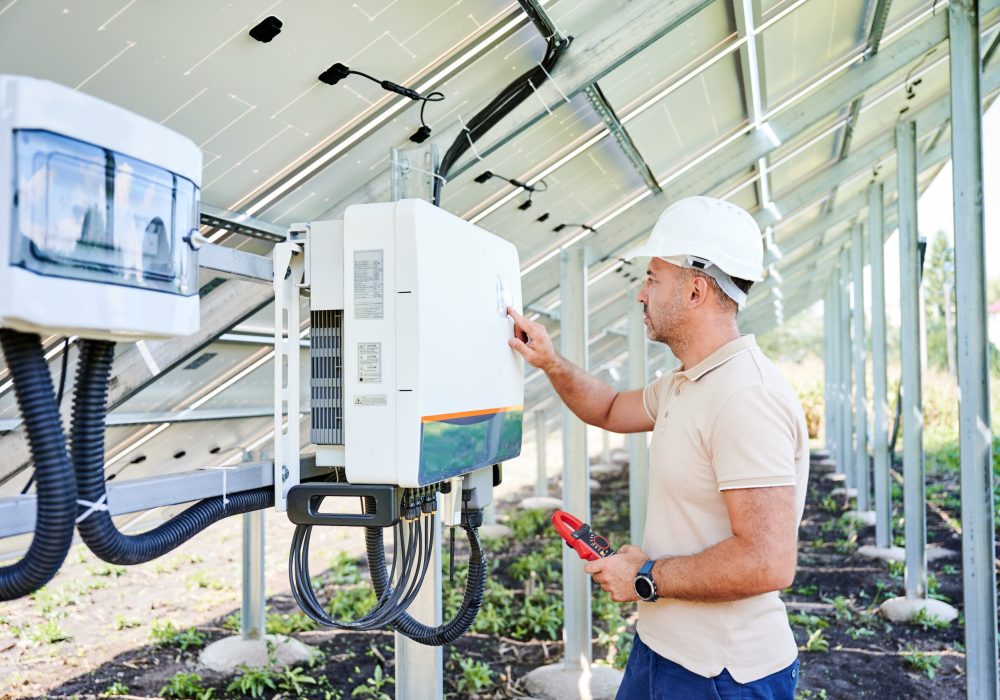 Professional worker checking voltage on solar inverter. Male using current probe to measure output voltage. Man in helmet pointing at inveter dial on back side of PV panel.